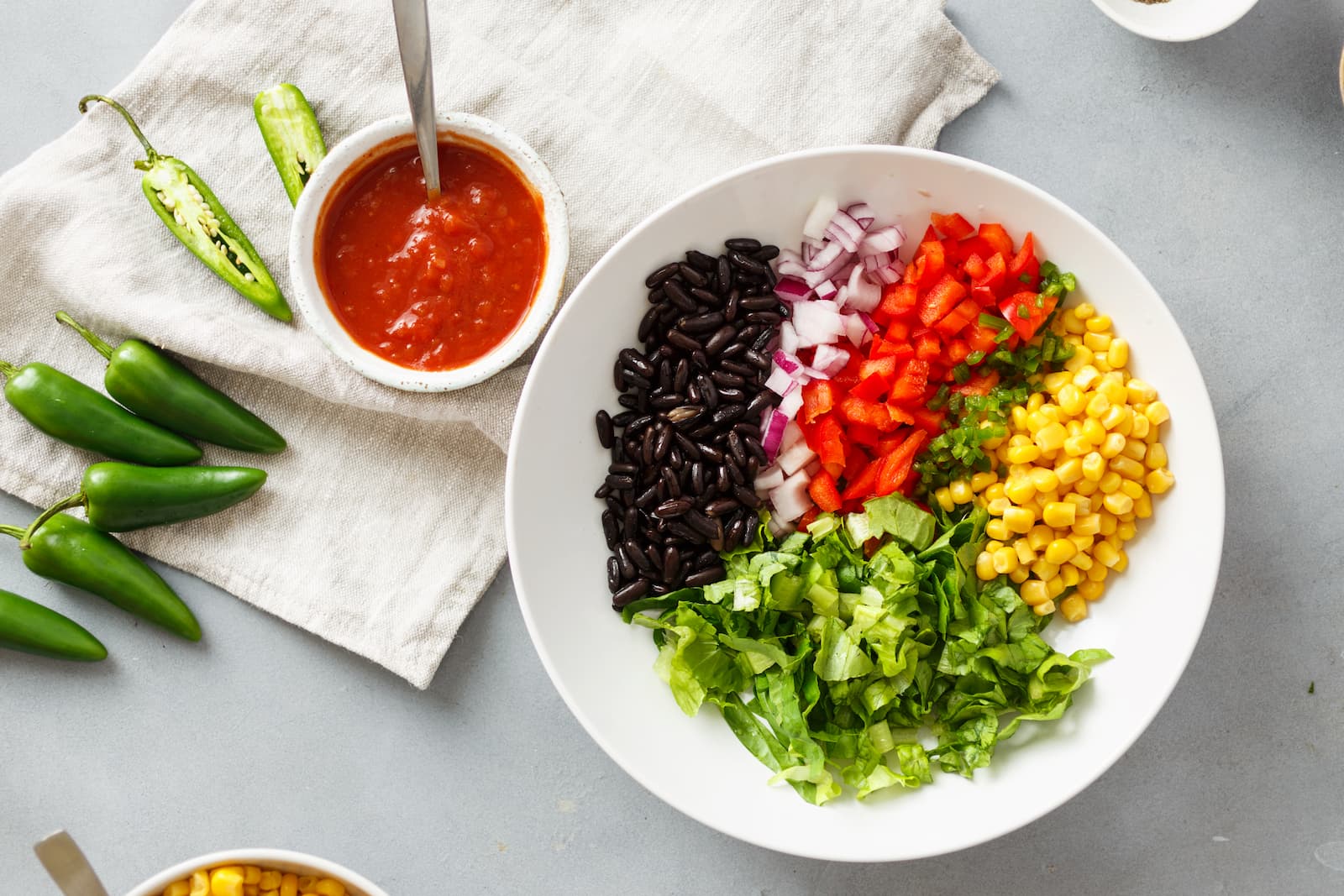 An overhead view of a large white bowl filled with neat sections of black beans, diced red onions, red bell peppers, chopped jalapeños, sweet corn, and shredded romaine lettuce, served alongside fresh salsa and whole jalapeños.